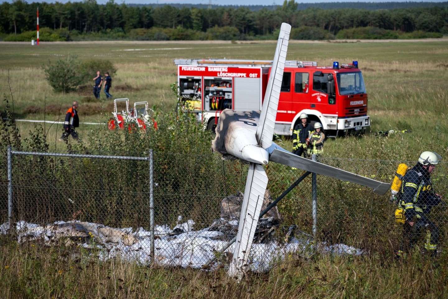 Trümmerteile eines Flugzeugs hängen in einem Zaun, während Einsatzkräfte der Feuerwehr an der Absturzstelle stehen.